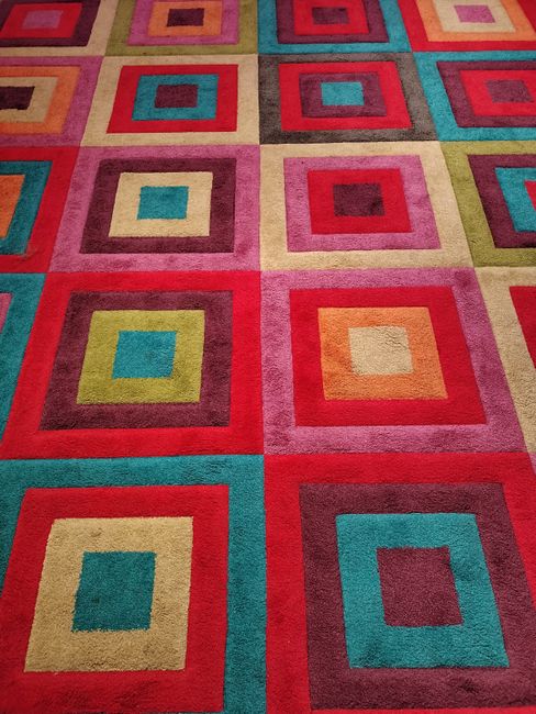 A colorful geometric-patterned rug featuring squares in various shades including red, pink, blue, green, beige, and orange, laid on a flat surface in a well-lit indoor space. The rug's surface appears